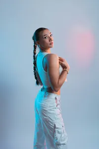 A woman with a long braid looks over her shoulder while illuminated by artificial blue and pink studio lighting, suggesting a futuristic and digital persona.